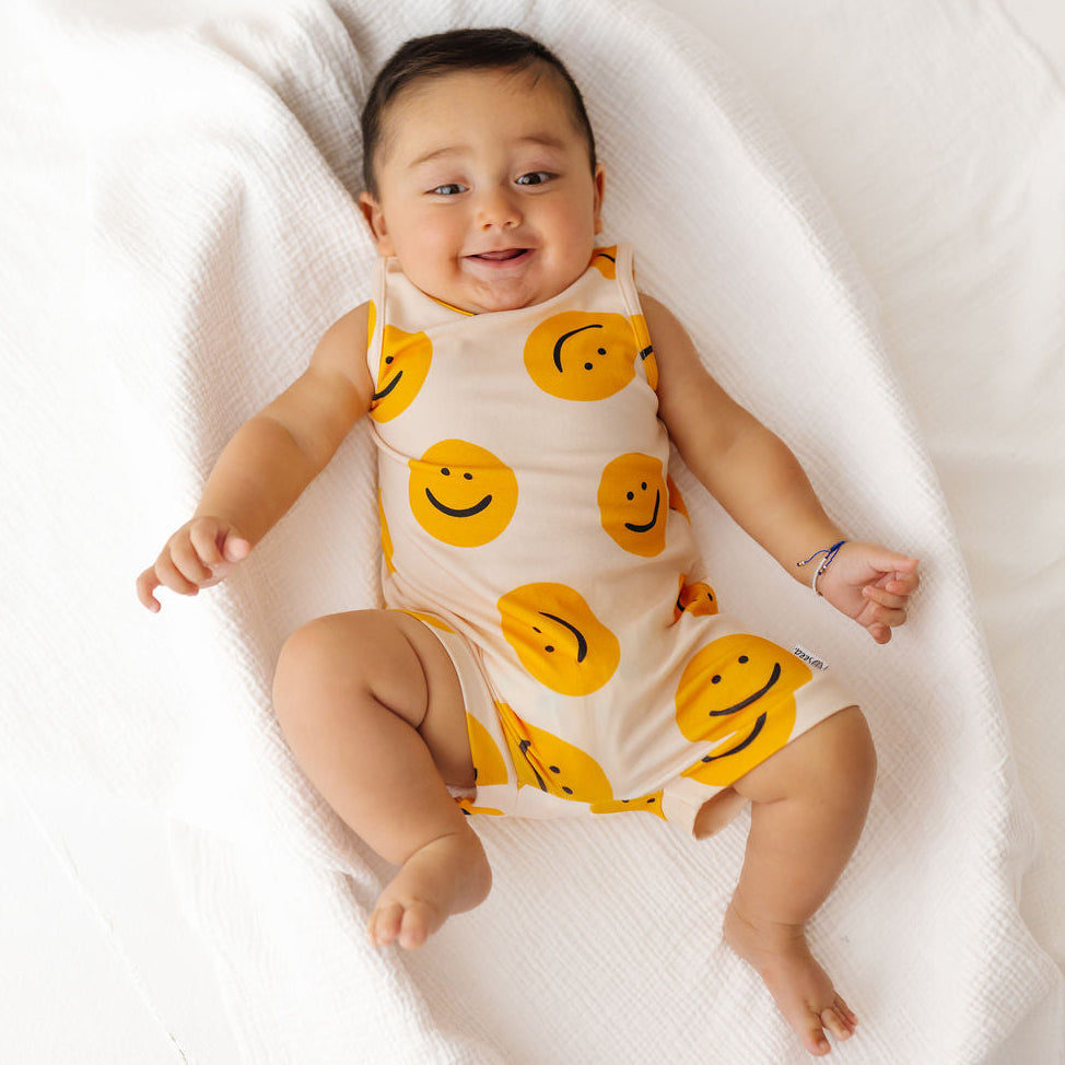Baby wearing a white onesie with yellow smiley face patterns, lying on a white blanket.