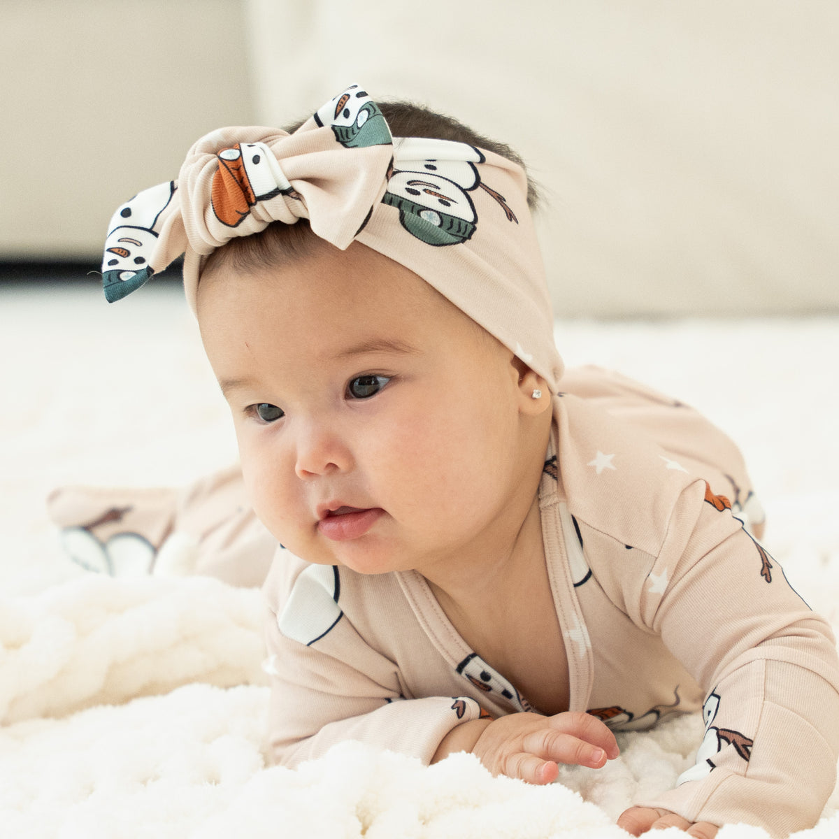 A baby lying on a white textured blanket, wearing a beige long-sleeve romper and a matching headband with a cartoon animal print.