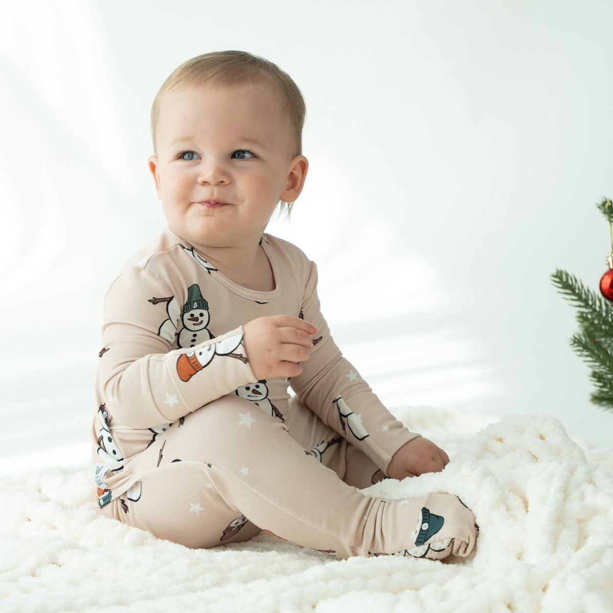 A baby sitting cross-legged on a white textured blanket, wearing a beige long-sleeve footed pajama with a cartoon snowman print. A small Christmas tree is visible on the right side of the frame.