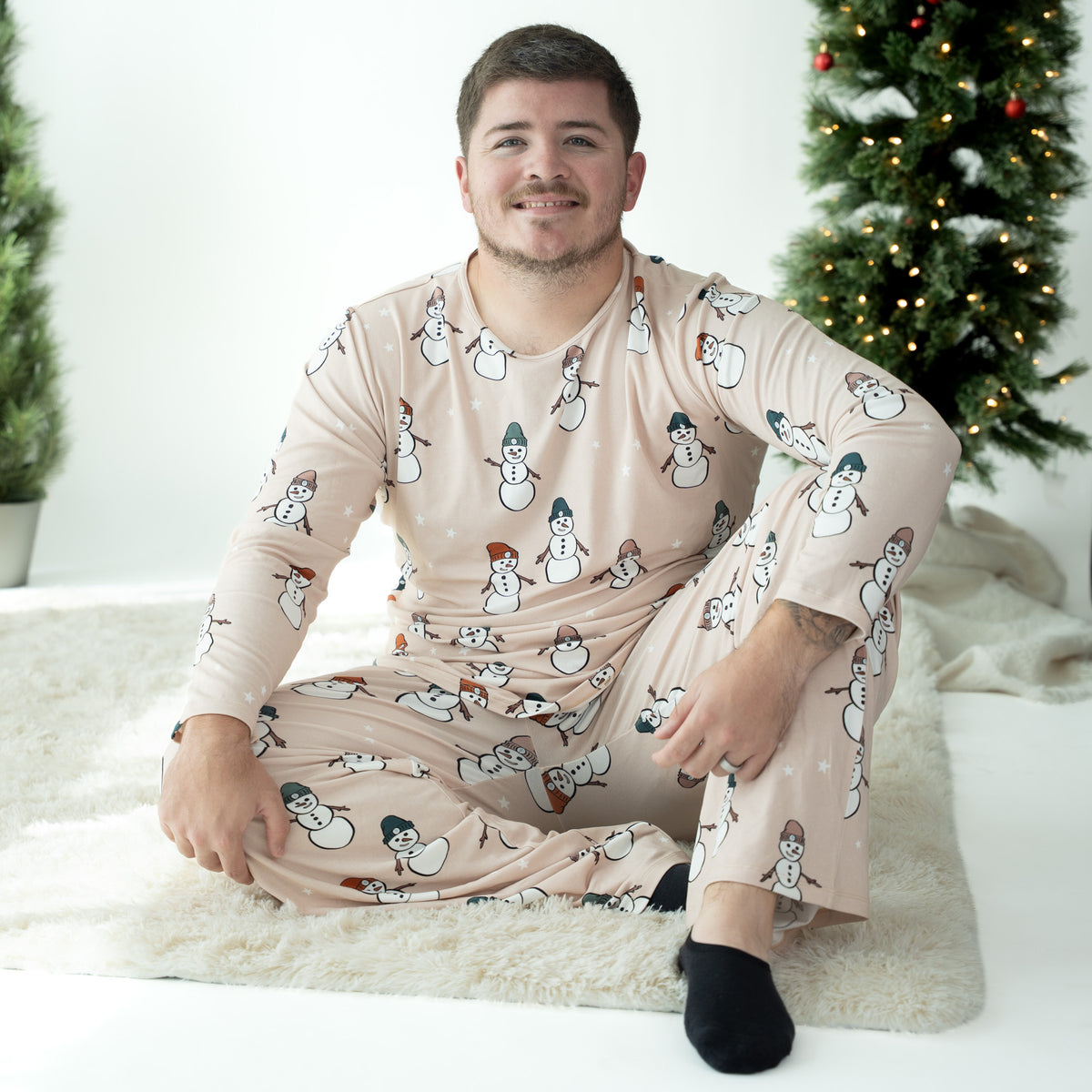 A man wearing a light beige long-sleeve pajama set with a snowman pattern, sitting on a white rug in front of a decorated Christmas tree.