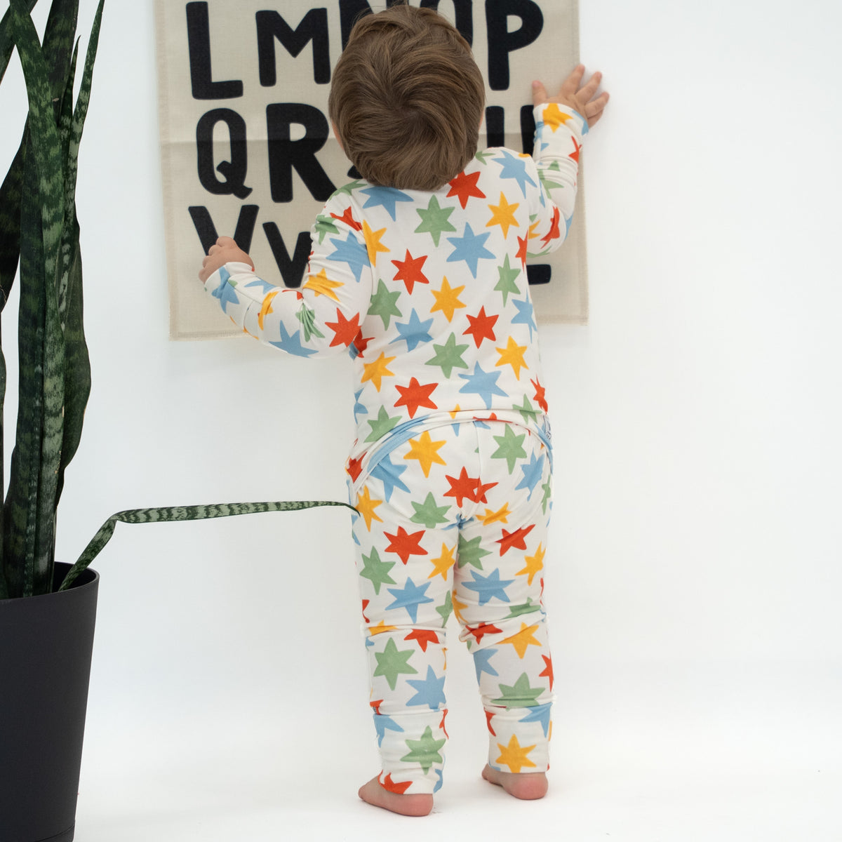 A toddler stands with their back to the camera wearing a cream-colored footed pajama set covered in a colorful pattern of red, orange, yellow, green, and blue stars. The child is posed in a bright room next to a potted snake plant, with a letter poster on the wall behind them.