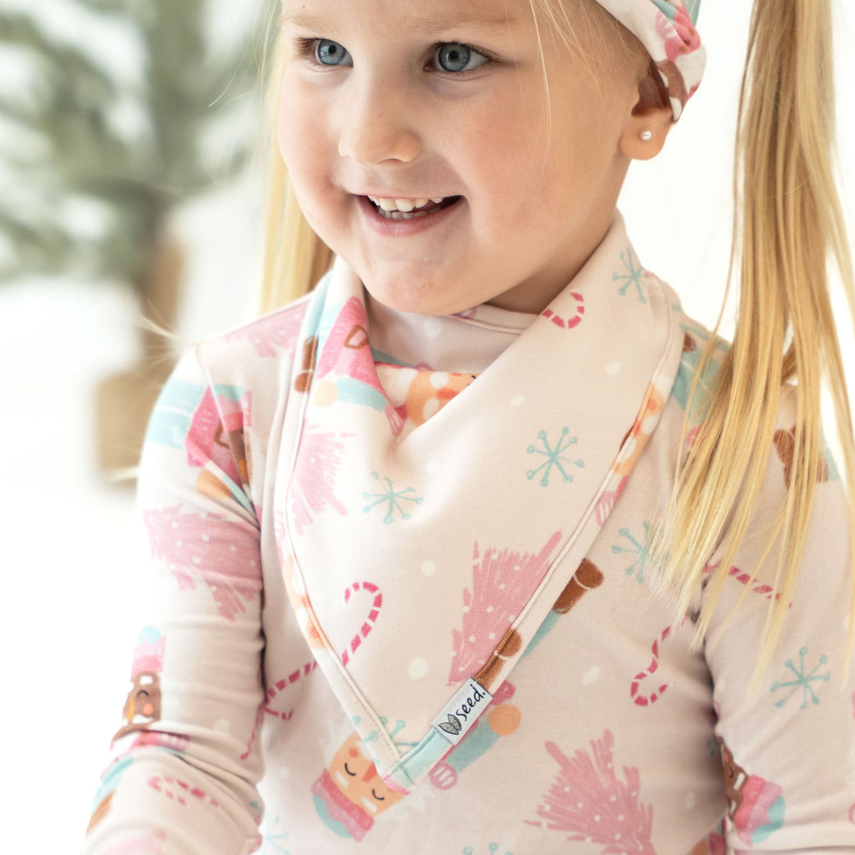 A close-up of a young girl wearing a soft cream-colored bib with a colorful winter print of gingerbread cookies, candy canes, and snowflakes, paired with a matching long-sleeve top and a patterned headband.