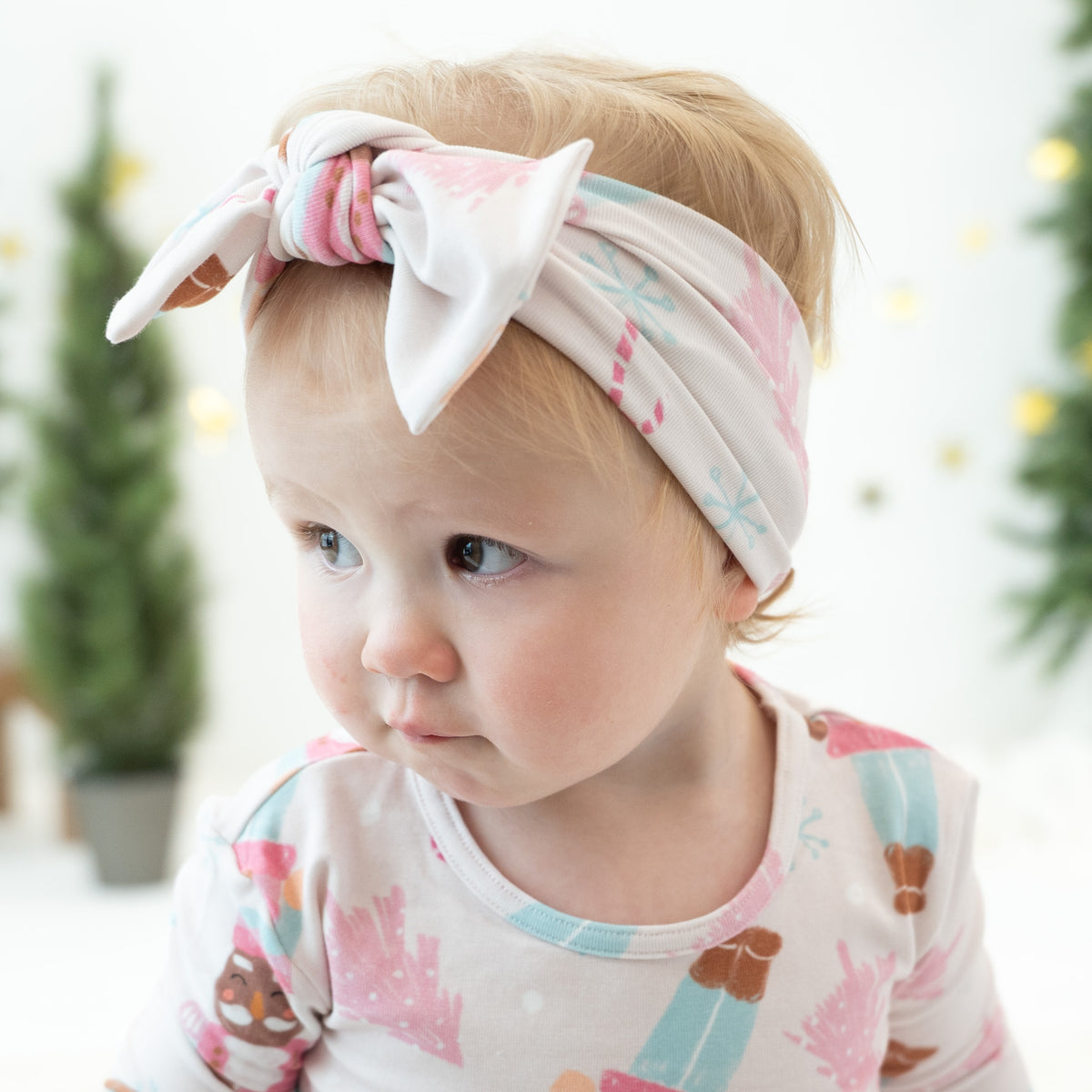 A baby wearing a light pink and white patterned fabric headband with a large bow, paired with a matching printed top. The background is a soft, bright white with blurred green trees.