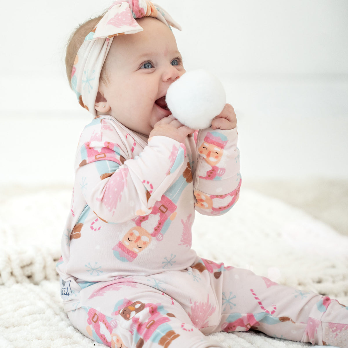 A baby wearing a light pink long-sleeve footed sleeper with a colorful holiday nutcracker print and a matching bow headband, sitting on a white textured blanket and holding a white teething toy.