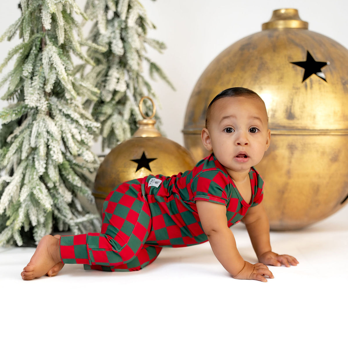 A baby crawling on a white studio floor wearing a red and green checkered holiday pajama romper, posed in front of a flocked Christmas tree and gold ornament props.
