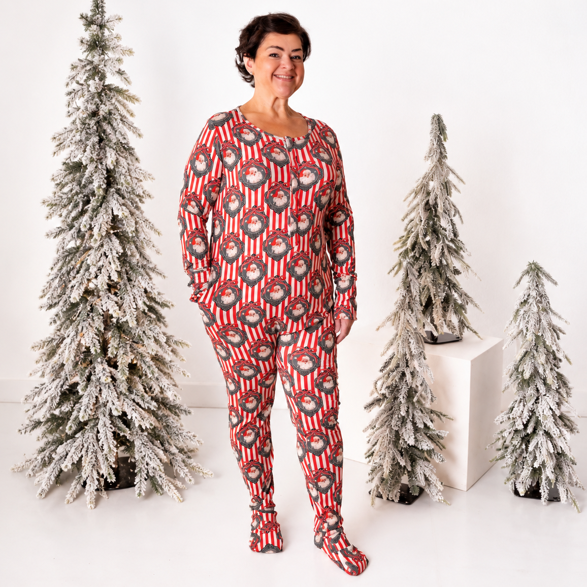 A woman stands in a studio wearing a red and gray holiday-themed two-piece pajama set with a flocked Christmas tree pattern. She is flanked by three artificial flocked Christmas trees on a white background.