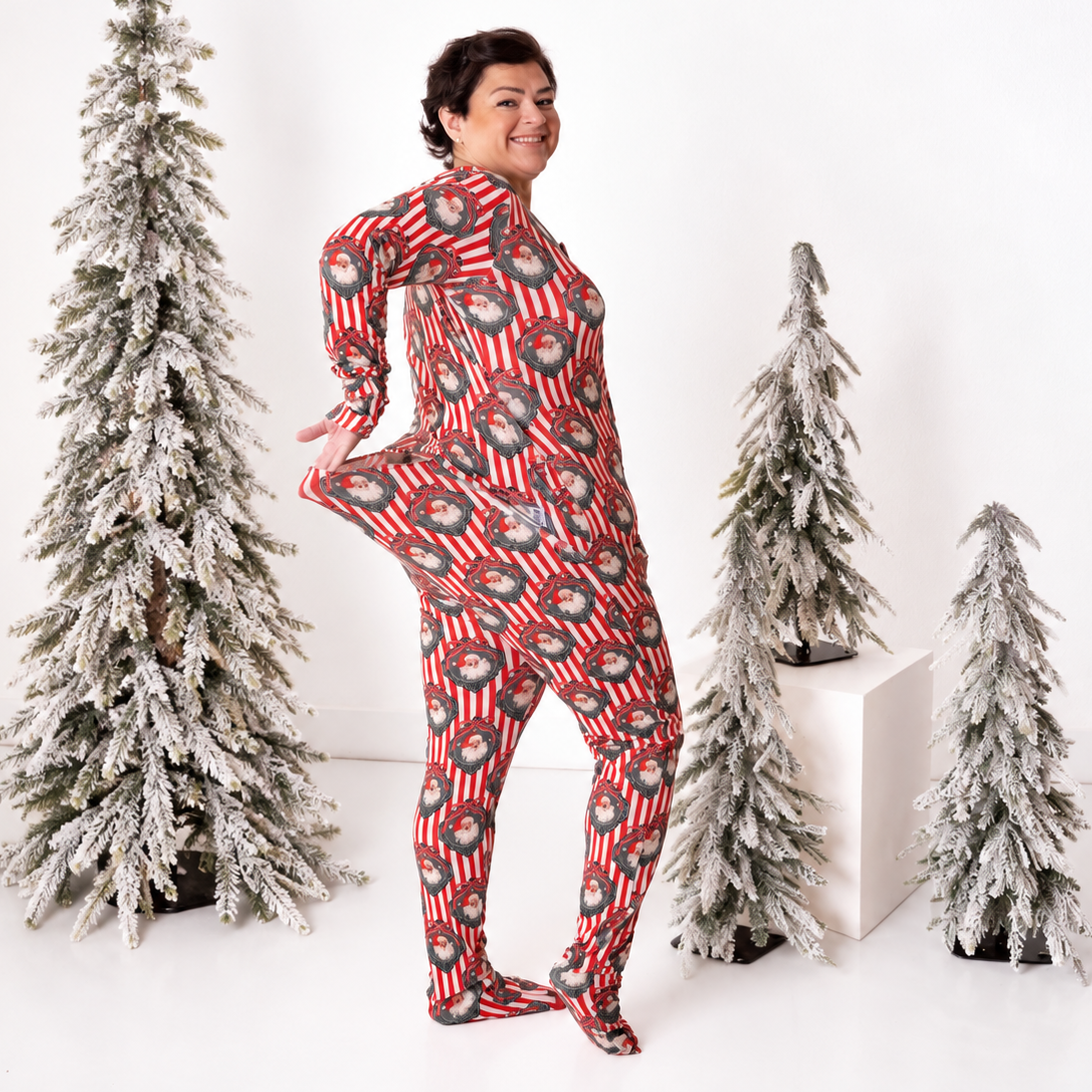 A woman wearing a red and white striped Christmas-themed full-body jumpsuit with a snow globe pattern, standing in a studio with three flocked Christmas trees.
