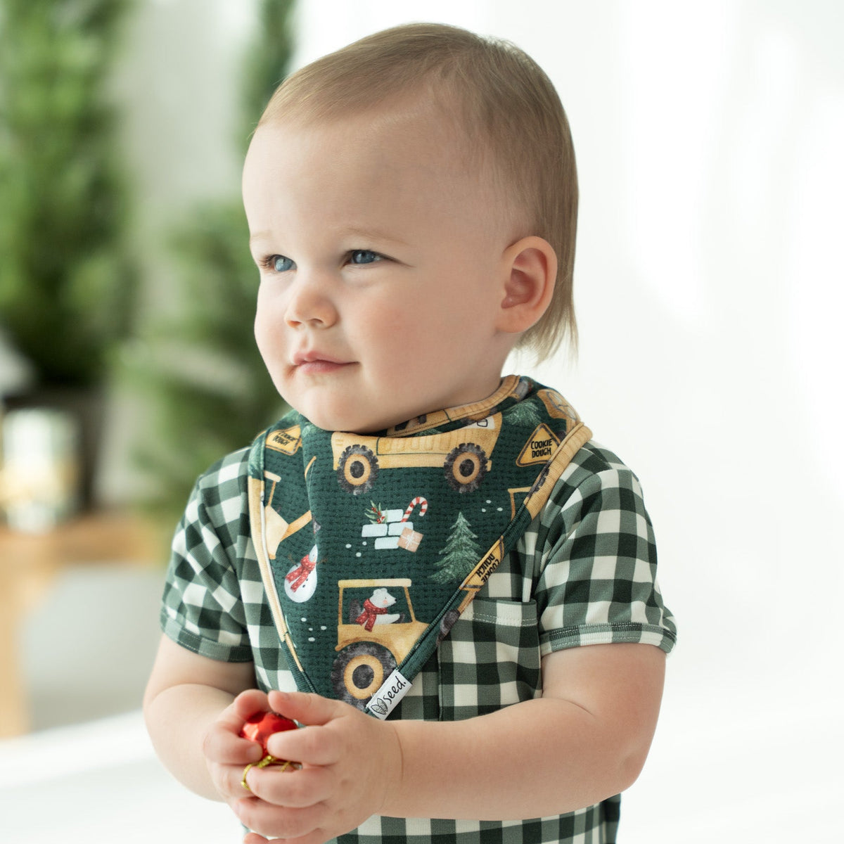 A baby wearing a green and white gingham romper and a dark green bib with a construction worker and Christmas tree pattern, standing in a bright indoor setting.