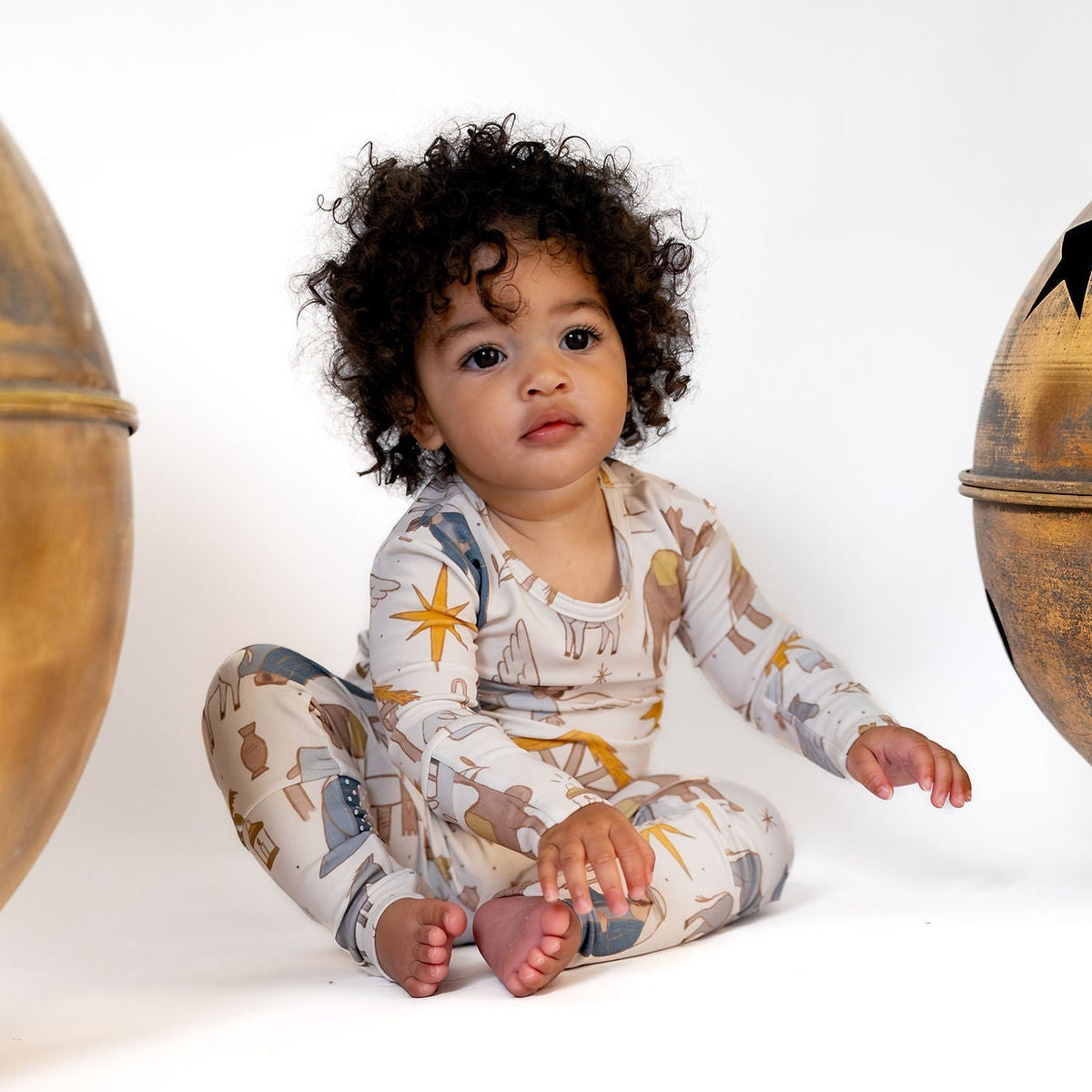 A baby with curly dark hair sits cross-legged on a white studio floor, wearing a long-sleeve one-piece pajama with a celestial print. The pajama has a cream base with gold, blue, and brown star and moon motifs.