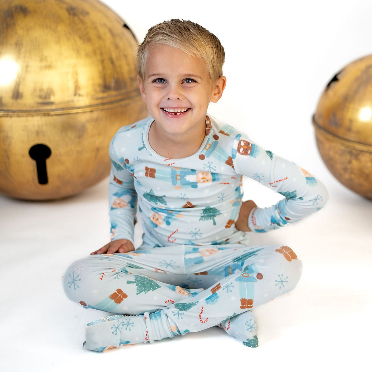 A young boy sits cross-legged on a white studio floor wearing light blue Christmas-themed pajamas with a print of gingerbread houses, trees, and candy canes. The background features large decorative gold bells and a small decorated tree.