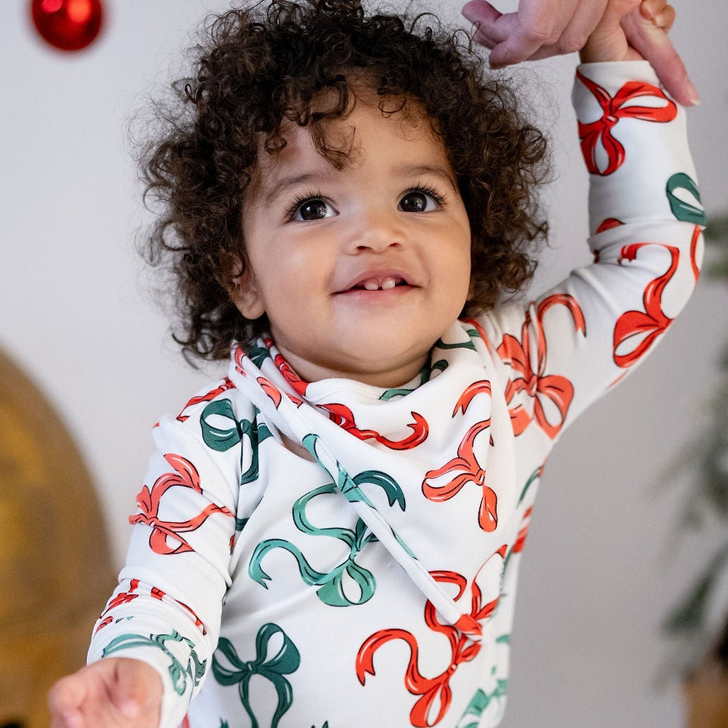 A toddler wearing a white long-sleeve bodysuit with a red and green ornament print, standing barefoot in a festive studio setting with large gold bells and holiday decorations.