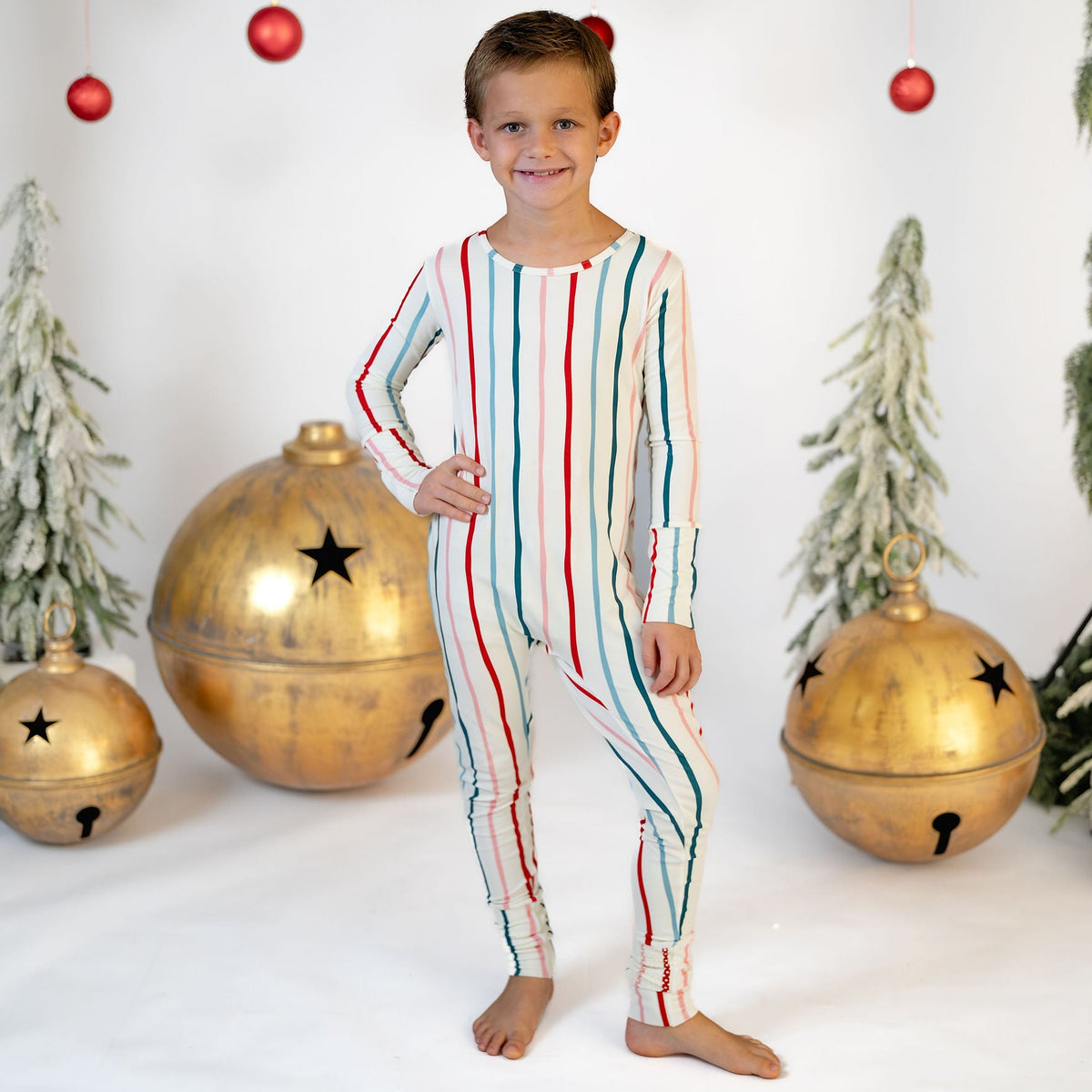 A young boy stands barefoot in a holiday-themed studio setting, wearing a long-sleeve one-piece pajama with vertical stripes in red, green, and blue on an off-white base. The background features a white wall decorated with hanging red ornaments, artificial pine trees, and large gold bells.