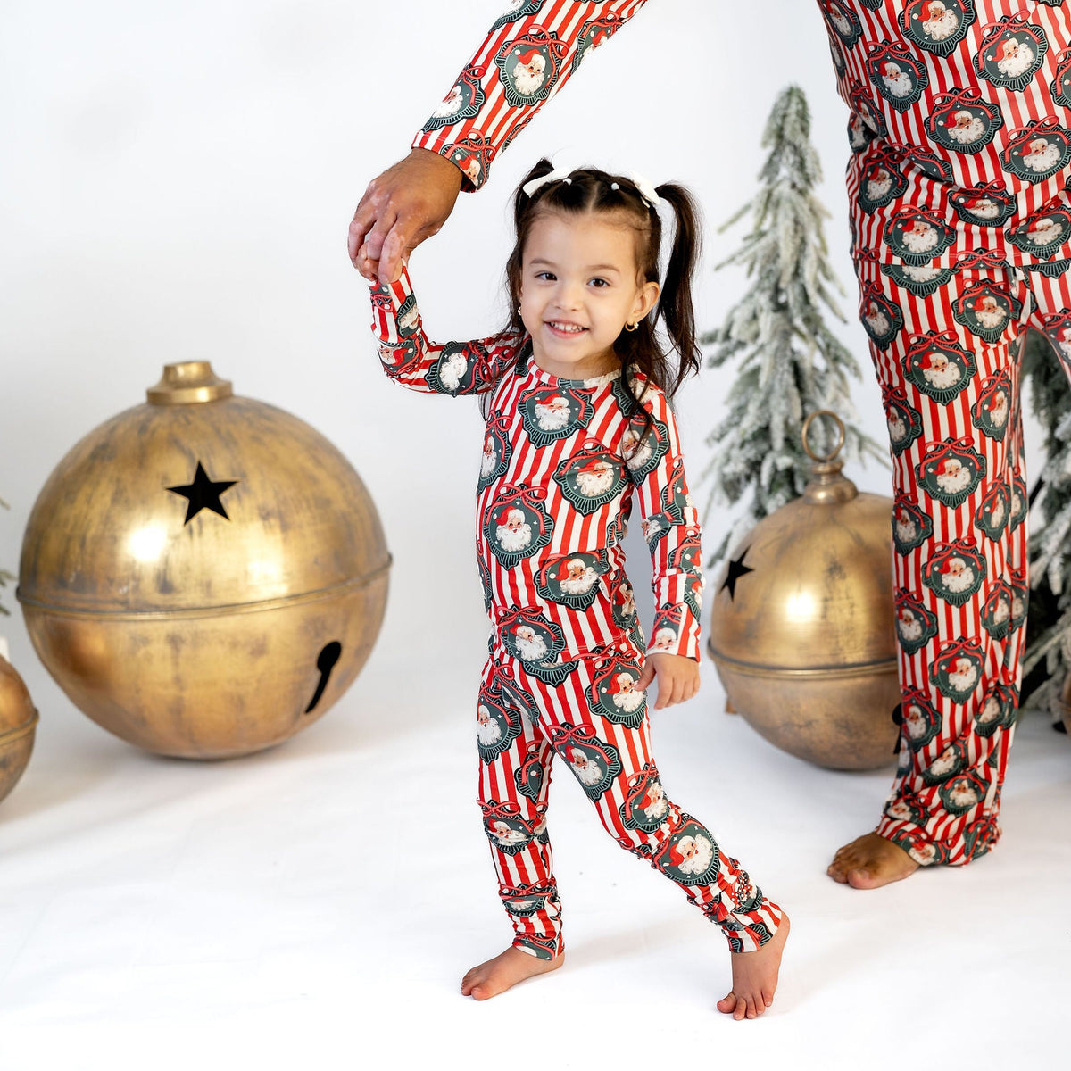 A young girl wearing a red and white Christmas-themed pajama set with a red-and-white striped background and green wreath patterns, walking barefoot while holding an adult's hand. The scene is decorated with miniature trees and large gold ornaments against a white studio background.