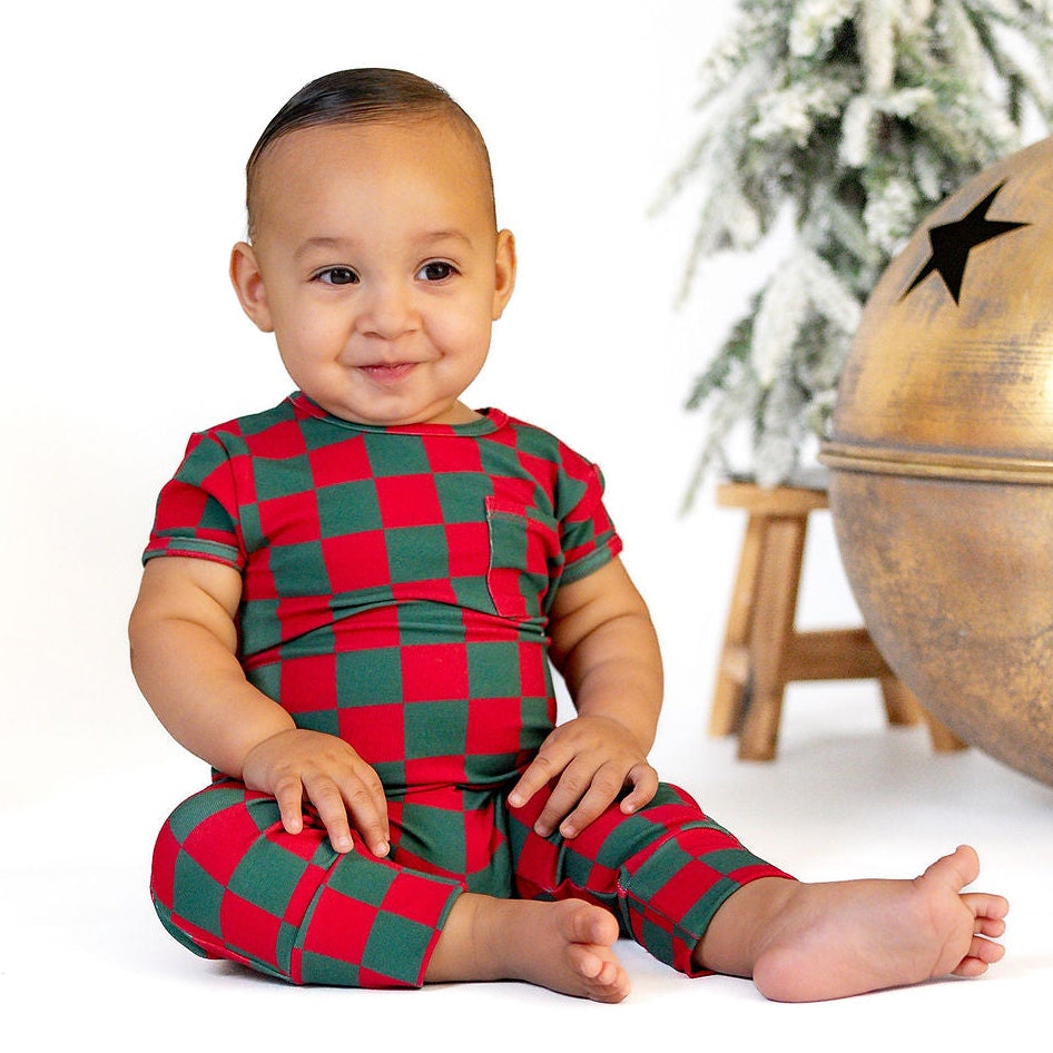 A baby wearing a red and green checkered holiday pajama set sits on a white studio floor between two large gold bell ornaments, with a small decorated tree in the background.