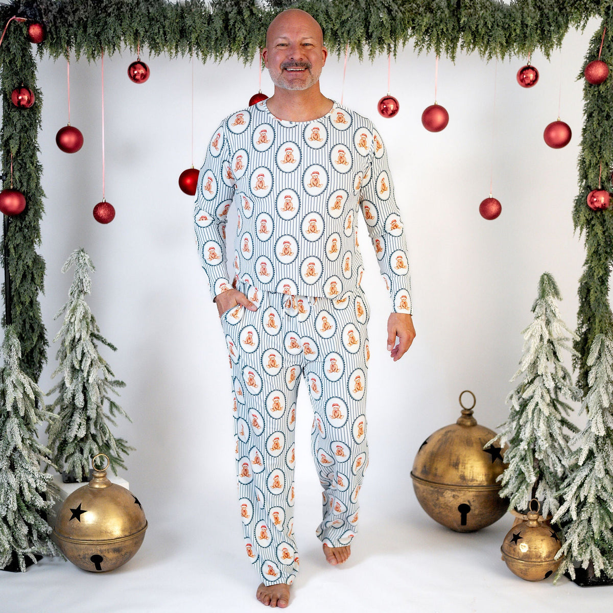 A man stands barefoot in a festive studio setting wearing a long-sleeve pajama set with a light blue background and an orange and white circular pattern. The scene is decorated with a green garland, red hanging ornaments, small flocked Christmas trees, and gold bells.