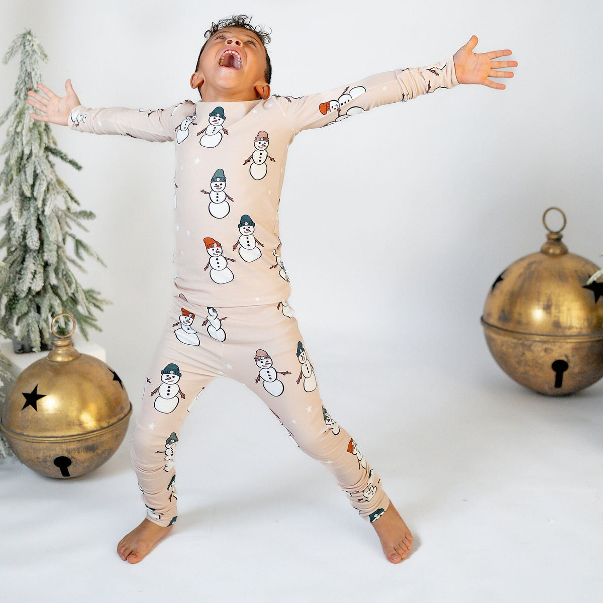A child wearing a beige long-sleeve pajama set with a snowman print, posing with arms outstretched in a festive holiday setting with miniature trees and gold ornaments.