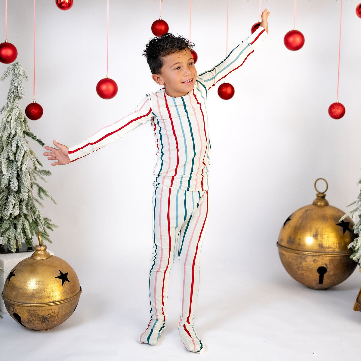 A young boy wearing a festive holiday jumpsuit with red, green, and white vertical stripes, posing with arms outstretched in front of a white studio backdrop decorated with Christmas garlands, red ornaments, and gold bells.