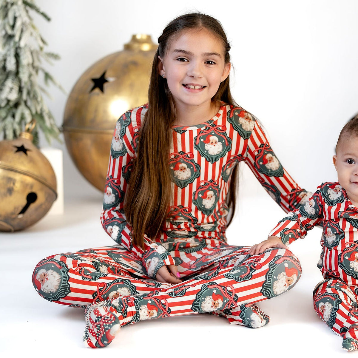 Two children wearing matching red and green Christmas-themed footed pajamas, sitting on a white studio floor with decorative gold bells and artificial snowy trees in the background.