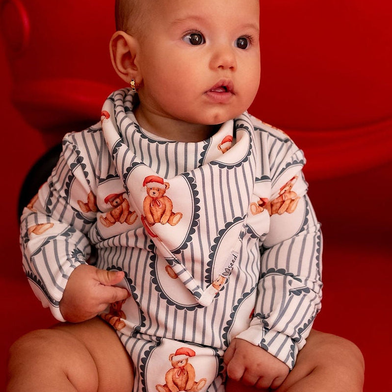 A baby wearing a white and gray pinstripe romper with orange cartoon characters sits in front of a red toy car decorated with a small evergreen branch, set against a solid red studio background.