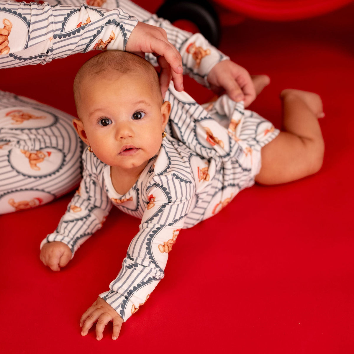 A baby wearing a white long-sleeve onesie with a navy blue wavy line pattern and orange cartoon motifs, crawling on a bright red floor while an adult in matching patterned pajamas adjusts the baby's shirt. A red toy cart is visible in the background.
