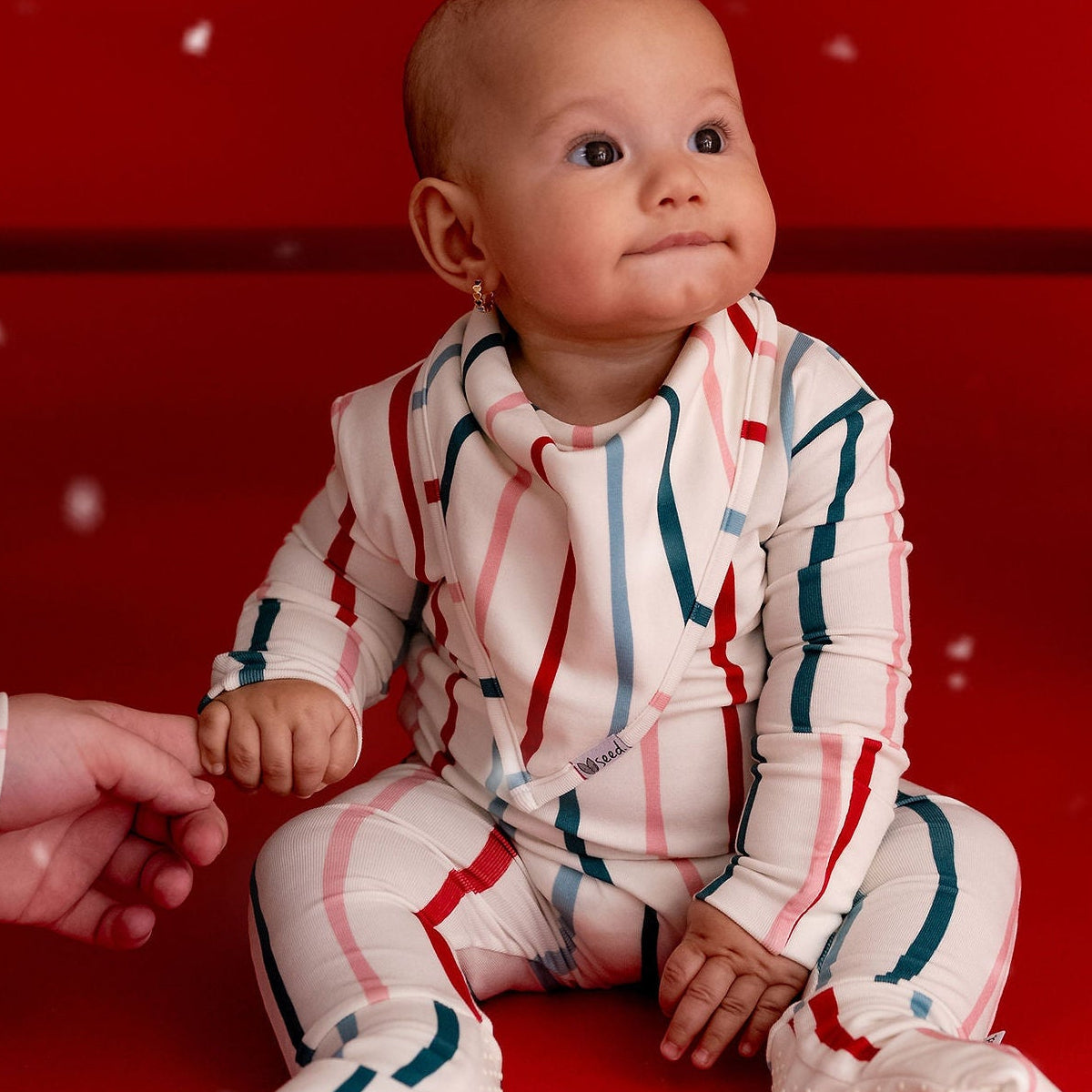 A baby wearing a festive red, white, and green striped knit pajama set sits on a red studio floor, holding an adult's hand. A red bench and a decorated Christmas tree are in the background with a snowfall effect.