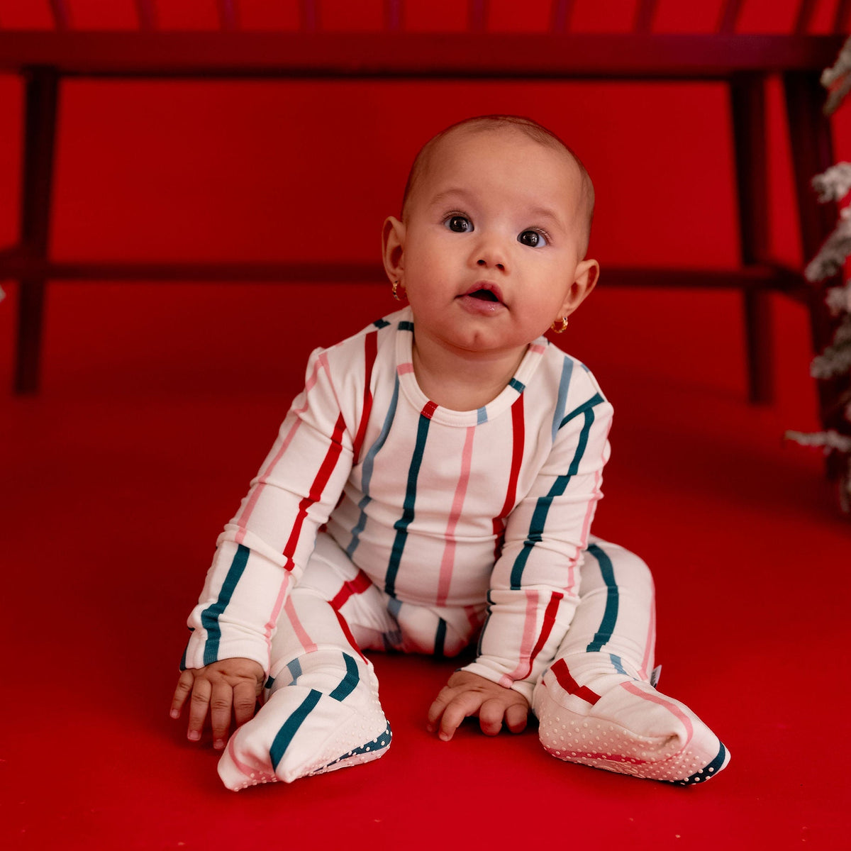 A baby sitting on a red studio floor wearing a white footed pajama set with red and teal stripes, posed in front of a dark wooden bench and red backdrop.