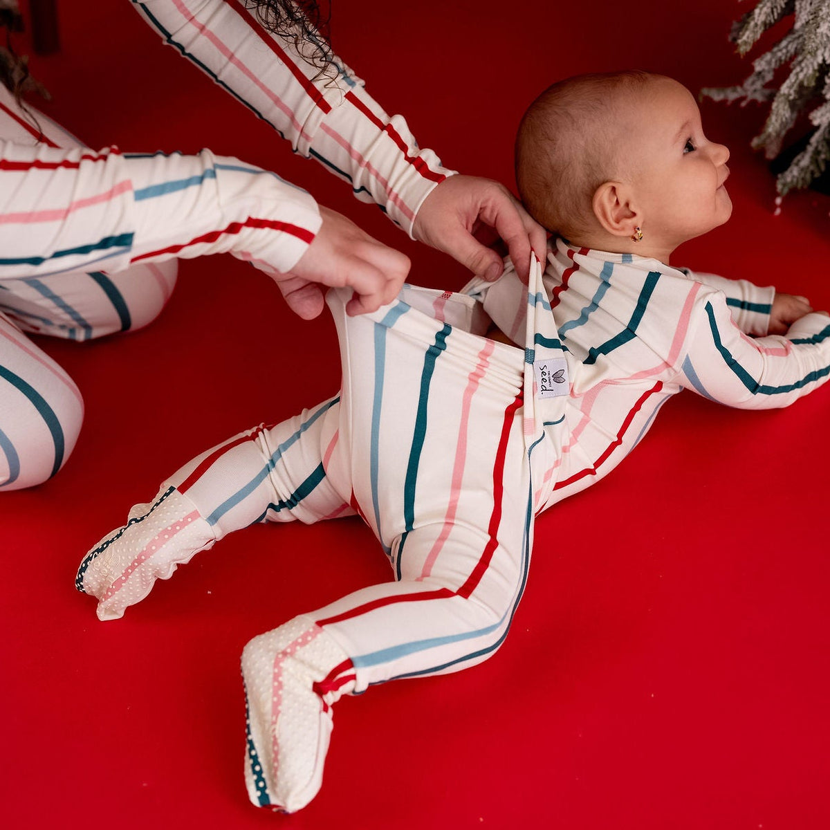 A baby wearing a red, white, and blue striped footed sleeper lies on a red floor while an adult adjusts the back zipper. A small flocked Christmas tree is visible in the upper right corner.