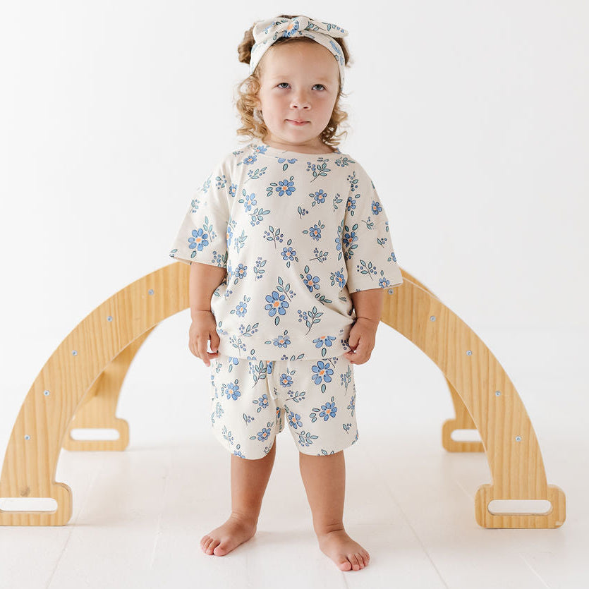 A toddler stands barefoot in front of a wooden arch play structure, wearing a matching cream two-piece outfit with a blue floral print and a coordinating floral headband.