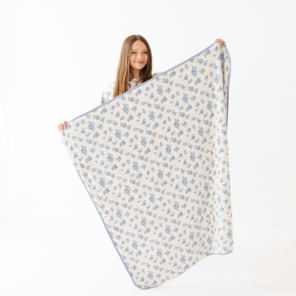 A young girl with long light brown hair holds up a large blanket or throw with a delicate blue floral pattern on a white background. She is standing against a seamless white studio background.