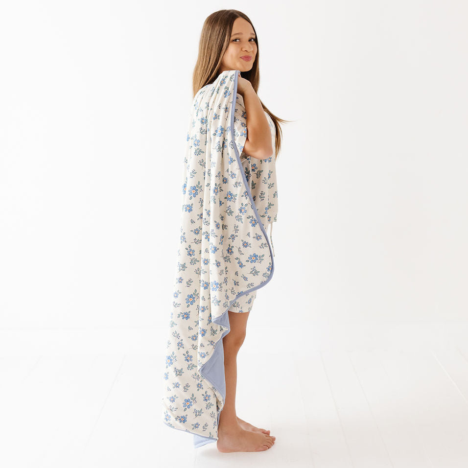 A young girl with long light brown hair stands in profile wrapped in a cream-colored cotton blanket with a blue floral pattern and a solid light blue border, set against a seamless white studio background.