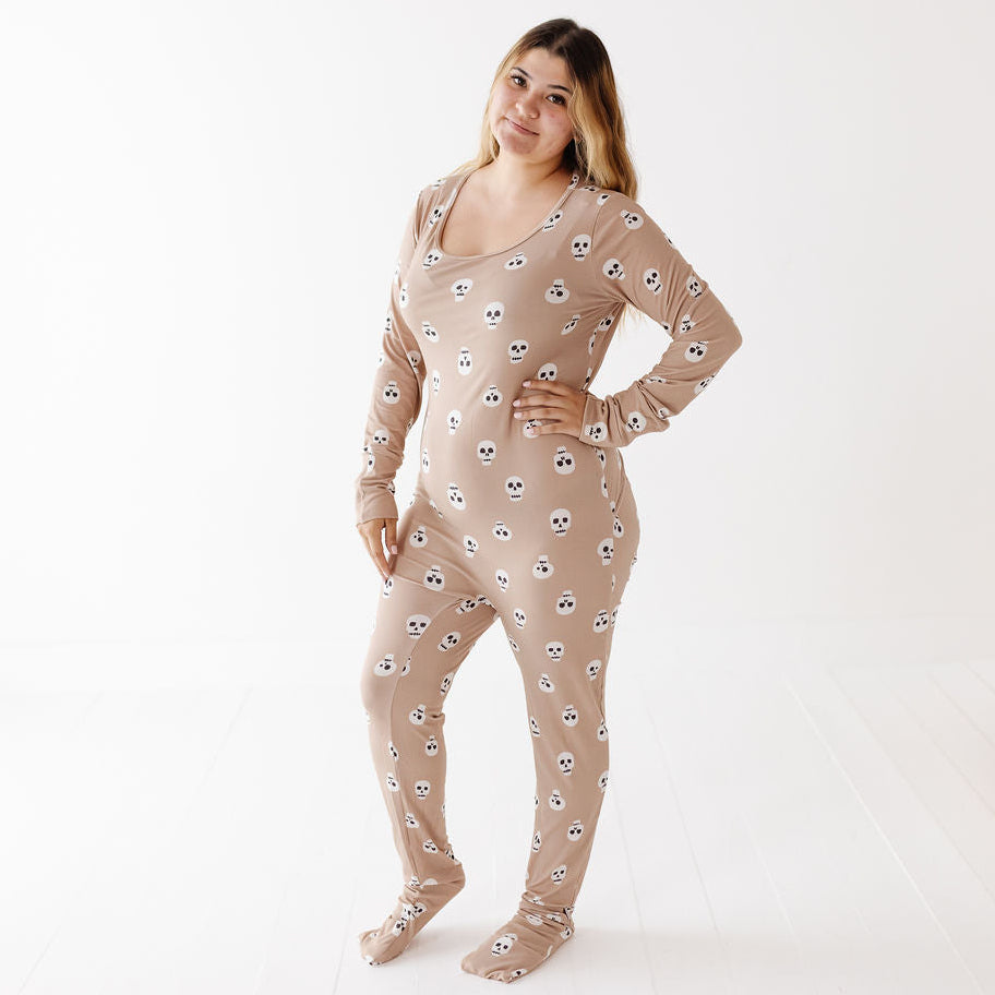 A woman modeling a beige long-sleeve footed pajama suit with a repeating skull and crossbones pattern, standing against a clean white studio background.