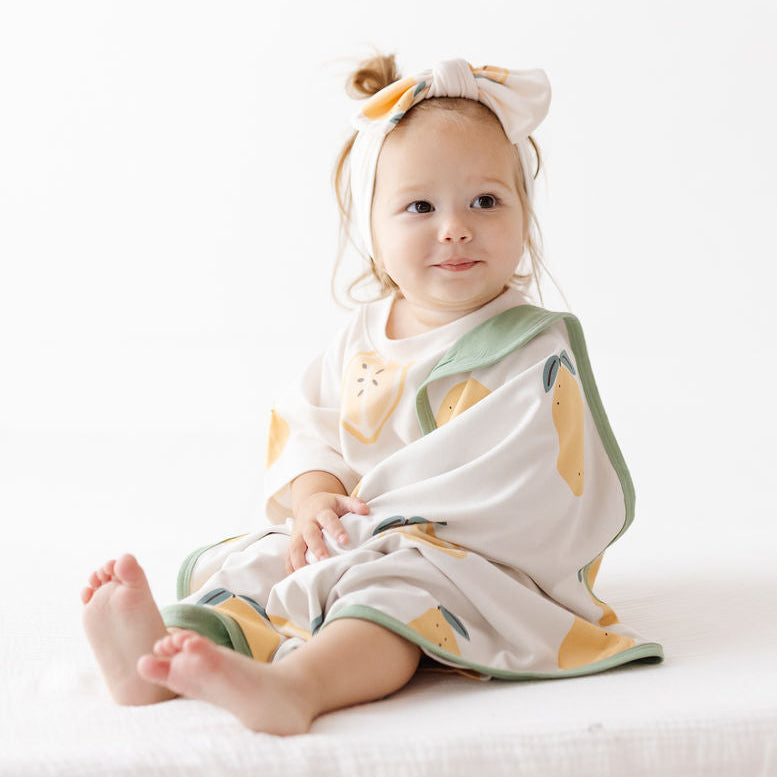 A baby girl sitting on a white fabric surface, wrapped in a white muslin blanket with a yellow and green citrus print, wearing a matching white bow headband.
