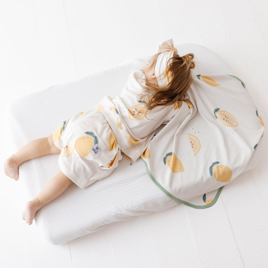 A baby lying on a white mattress, wrapped in a cream-colored blanket and wearing matching cream shorts with a yellow and green lemon print. A matching cream headband with a bow is also visible.
