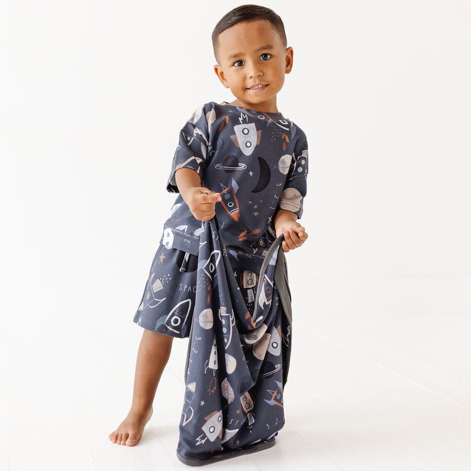 A young child wearing a dark navy blue cotton pajama set with a space-themed print of rockets, planets, and stars, standing against a plain white studio background.