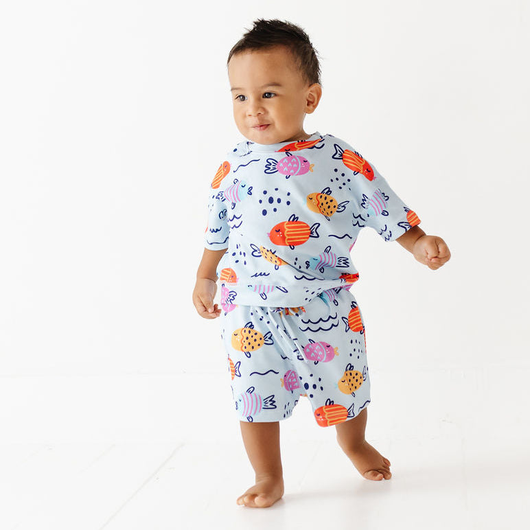 A toddler walking barefoot on a white studio floor, wearing a matching two-piece pajama set with an ocean fish pattern on a light blue background.