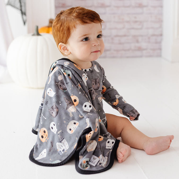 A baby with red hair sits on a white wooden floor wearing a gray hooded blanket with a Halloween print of ghosts, skulls, and pumpkins. The background features a light brick wall, white pumpkins, and white drapery.