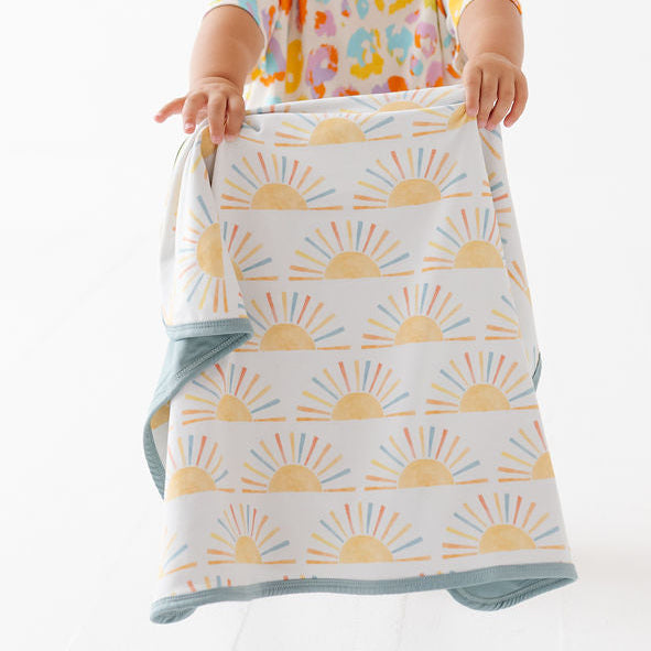A toddler wearing a colorful patterned shirt and a purple headband holds up a reusable cloth baby blanket with a yellow sunburst pattern and a blue border against a white studio background.