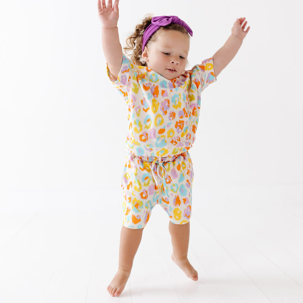 A toddler girl wearing a matching two-piece pajama set with a colorful abstract print, a purple headband, and barefoot on a white studio background.