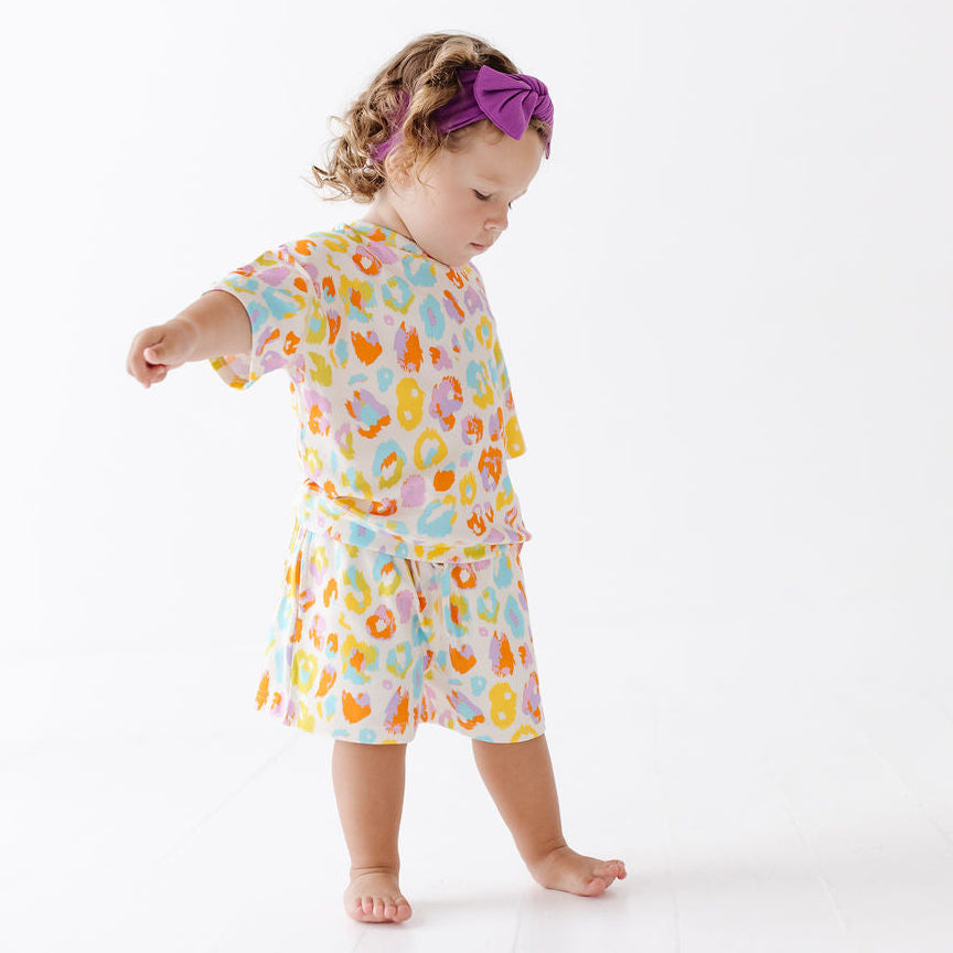 A toddler girl wearing a matching two-piece pajama set with a colorful abstract print, standing on a white studio floor. The set features a short-sleeve top and shorts in pastel shades of orange, yellow, pink, and blue on a white base.