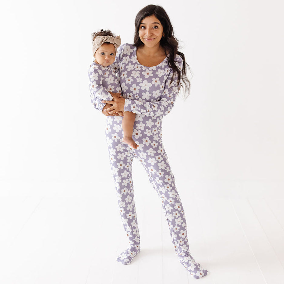 A woman holding a baby wearing matching long-sleeve footed pajamas with a white daisy floral print on a light lavender background, set against a clean white studio backdrop.