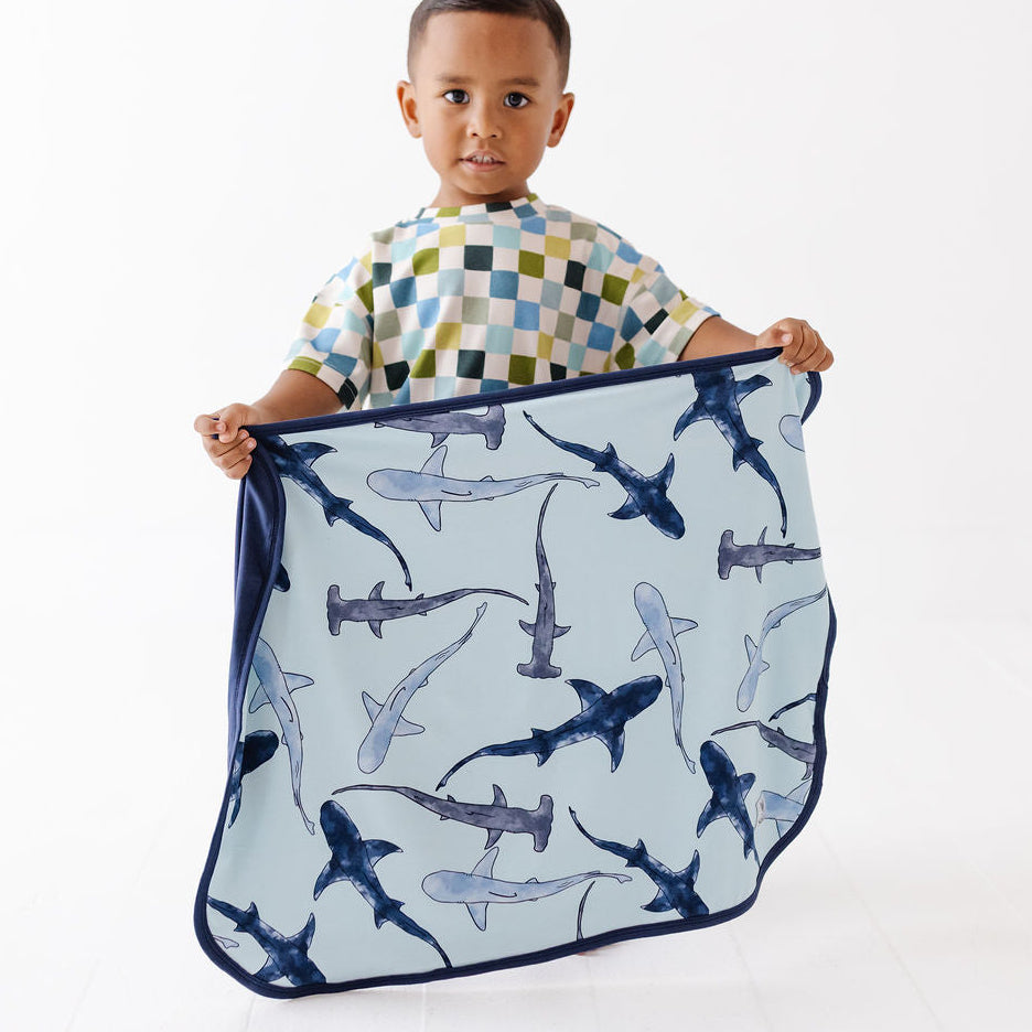 A young child stands against a white studio background holding up a square light blue blanket or towel with a dark blue shark print.