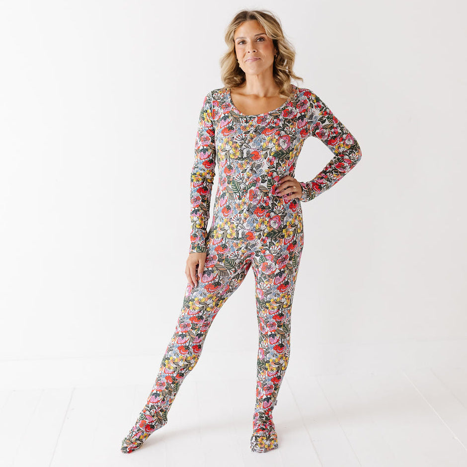 A woman modeling a long-sleeve, footed one-piece pajama with an all-over colorful floral and botanical print, posed against a clean white studio background.
