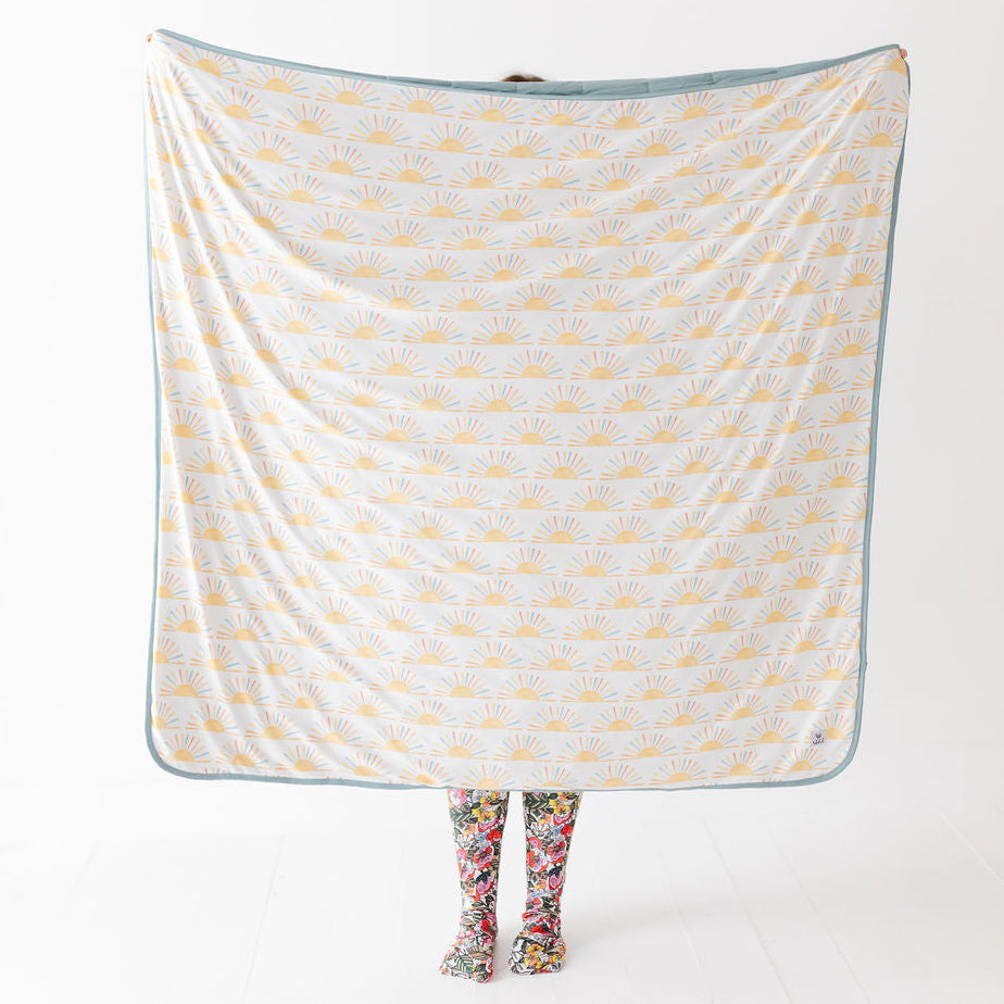 A person holds up a large square blanket or throw with a white background, yellow sunburst pattern, and a muted blue border. The person is only visible at the bottom through floral socks, against a clean white studio background.