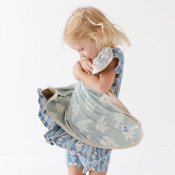 A young child stands barefoot against a clean white studio background, holding a large soft fabric bag or wrap in a light sage green color with a white leaf pattern. The child is also wearing a blue patterned dress.