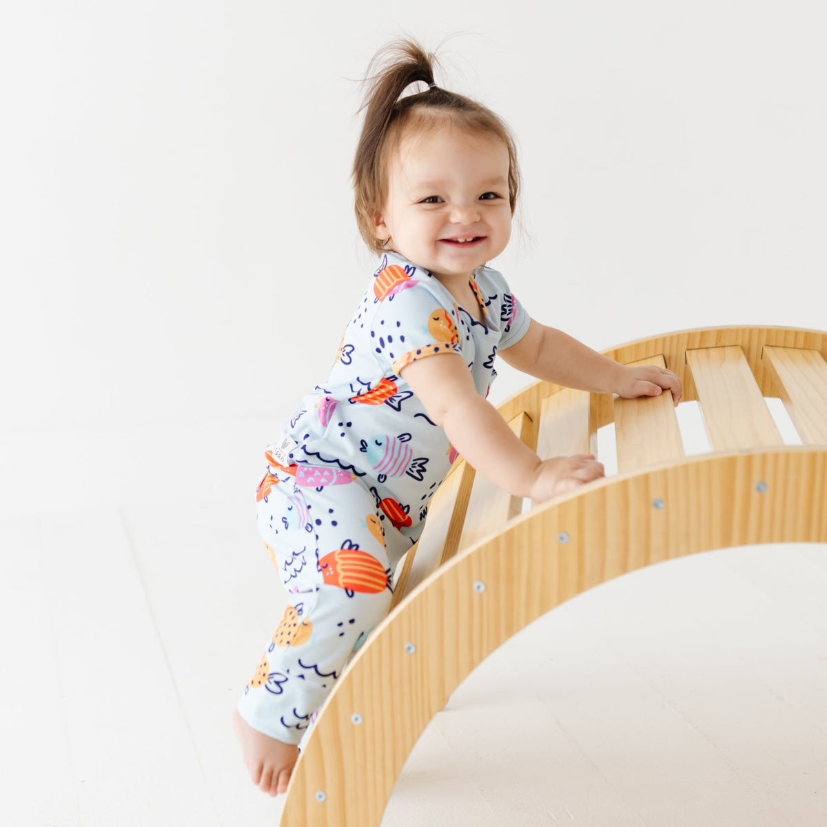 A smiling baby stands against a wooden arch on a white floor. She is wearing a light blue romped with a fish-themed print. 