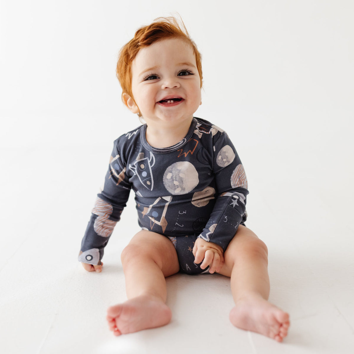 A smiling baby sits on a white floor wearing a navy long-sleeved onesie with a space-themed print. 