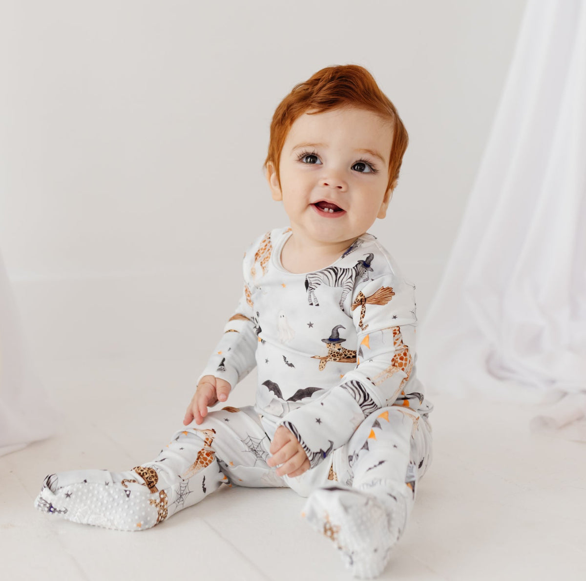 A smiling baby sits on a white floor, wearing a grey romper with a Halloween-themed print.