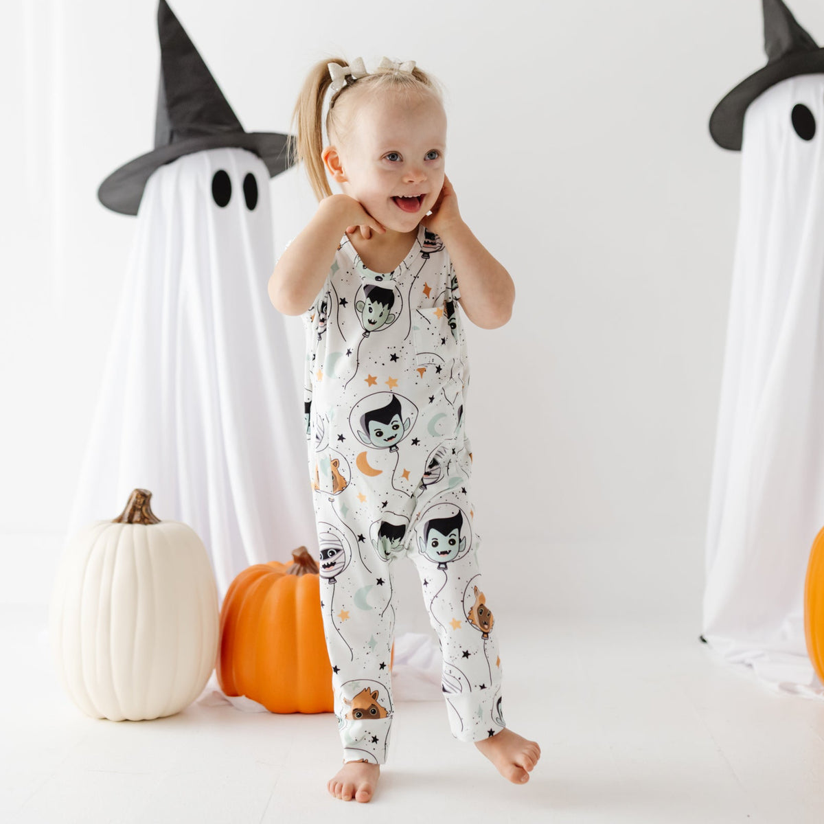 A smiling kid stand on white floor wearing white romper with Halloween design on it. 