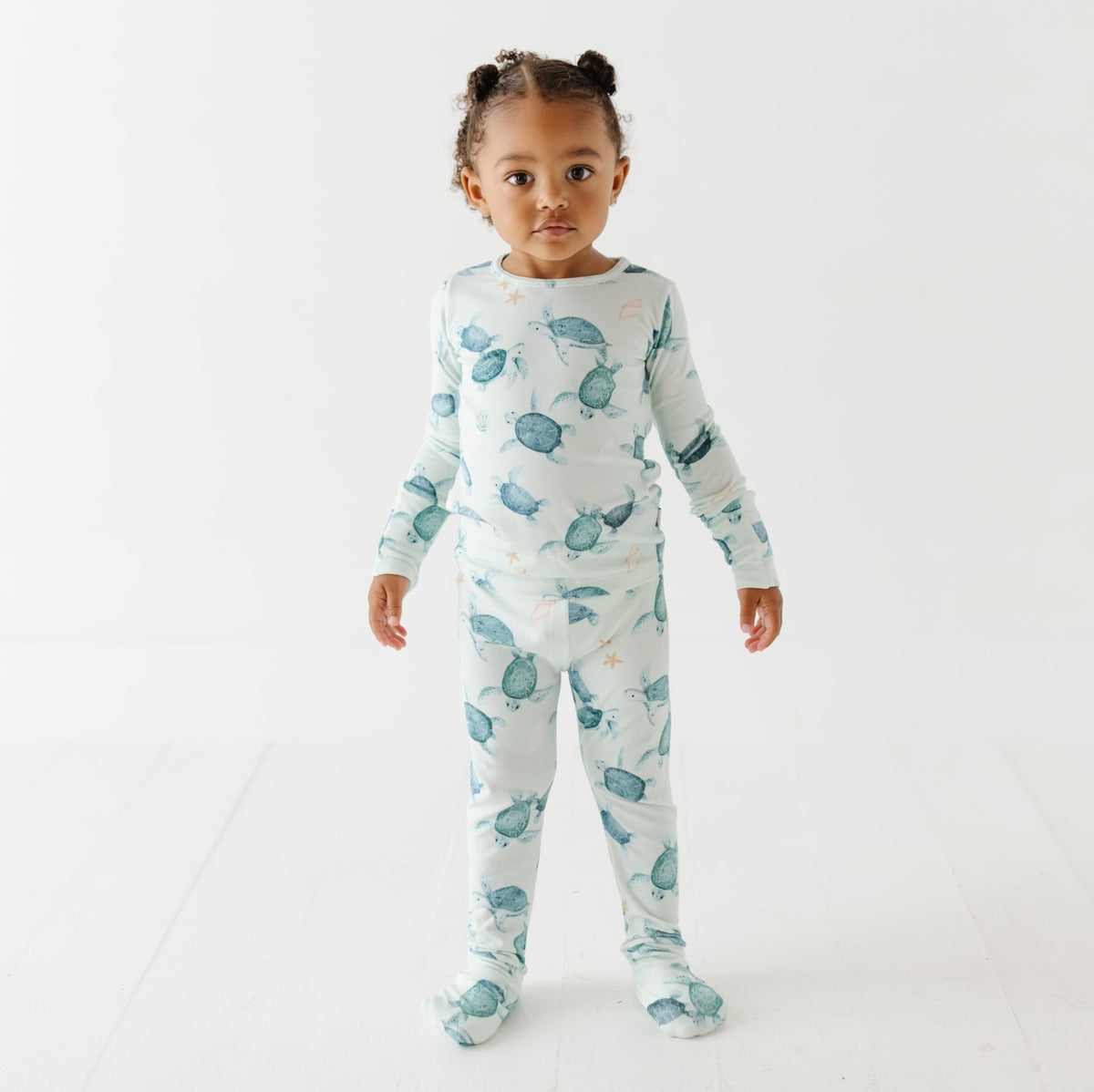 A kid stands on a white floor wearing a light green pajama set with turtles on it.