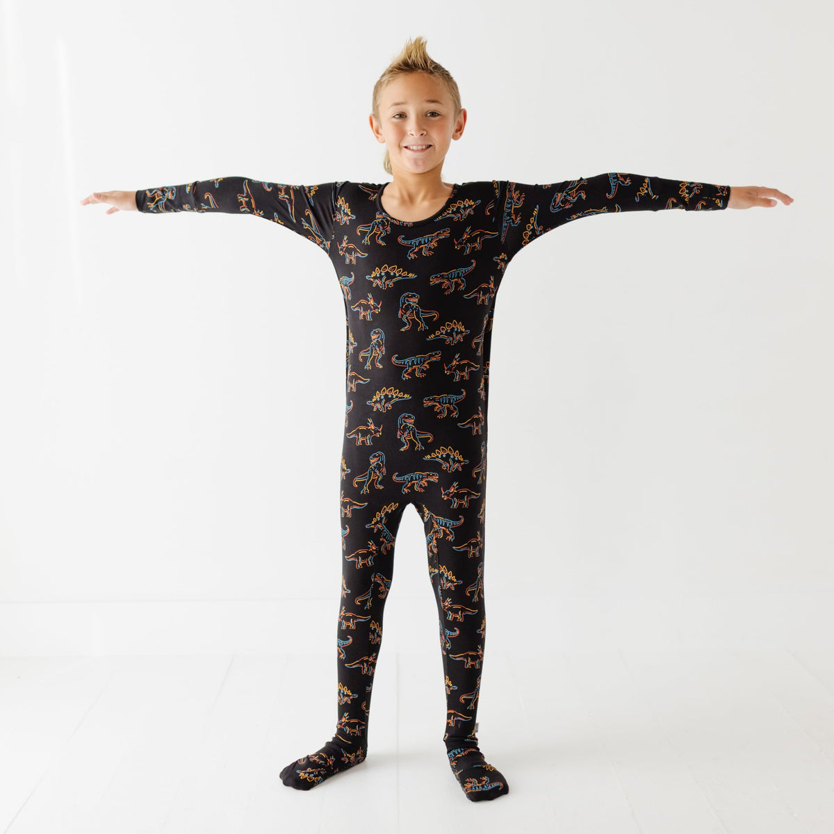 A kid stand on white floors wearing black romper  with glowing dinosaurs on it. 